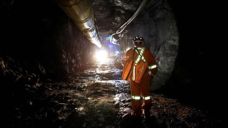 A miner, clad in full orange workwear, walks towards the entrance of a dark, underground tunnel.