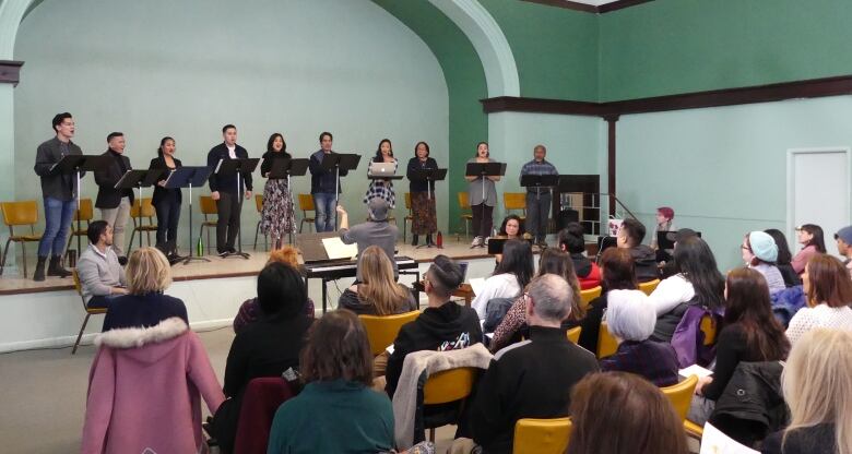 Ten people are pictured singing on stage for a group of people.