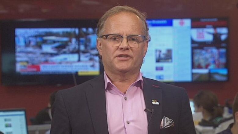 A man in a pink shirt, suit and glasses sits in a TV studio in front of multiple screens.