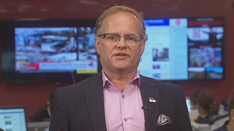 A man in a pink shirt, suit and glasses sits in a TV studio in front of multiple screens.