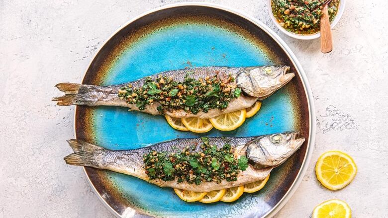 Overhead shot of 2 whole, roast sea bass topped with herbs. They're on a turquoise serving platter on a grey surface. 