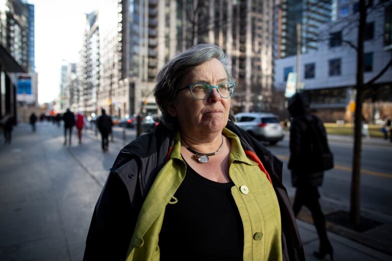 A woman with grey hair, blue glasses, a green shirt and a black jacket stands on the sidewalk of an urban street