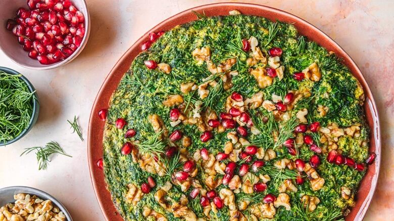 Overhead shot of a frittata with lots of herbs topped with fresh dill, pomegranate arils and walnuts. It's sitting on a pink serving platter on a pink table.  