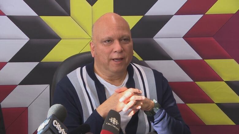 A bald man in a navy jacket with vertical black and grey stripes folds his hands as he speaks at a news conference. He's pictured behind microphones and against a quilted backdrop that's yellow, white, red and black.