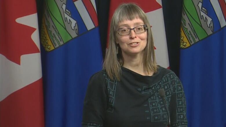 A woman in long hair and glasses stands in front of Canadian and Albertan flags.