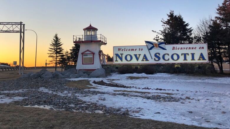 A sign reads Welcome to Nova Scotia in both English and French. There is snow on the ground and the sun is setting in the distance.