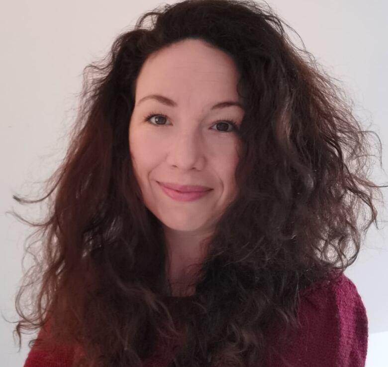 A woman with brown curly hair standing against a white wall with her arms crossed.