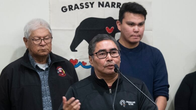 A man speaks into a microphone with two people behind him. Behind them is a sign that reads, 