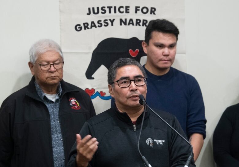 A man speaks into a microphone with two people behind him. Behind them is a sign that reads, 