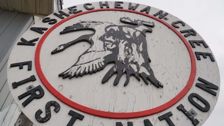 A circular white wooden sign with paint peeling off of it reads 'Kashechewan Cree First Nation' with a logo in the middle depicting a man's head, a goose and a moose.