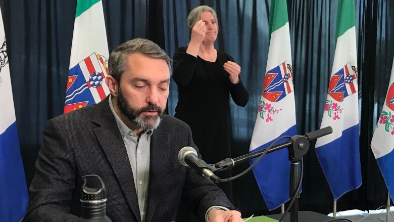 A man at a table sits before a microphone reading from some papers.