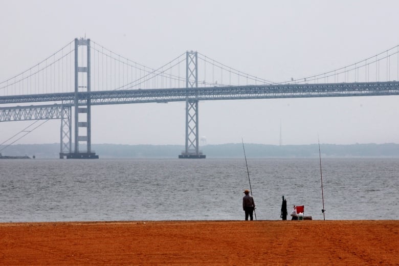 A man with a long fishing pole is in the foreground with a huge bridge spanning a body of water in the background. 