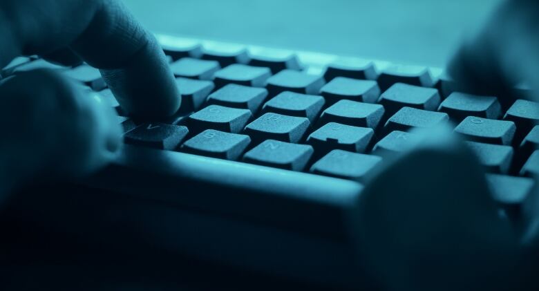 A closeup shows the hands of a person typing on a keyboard in a darkened room.