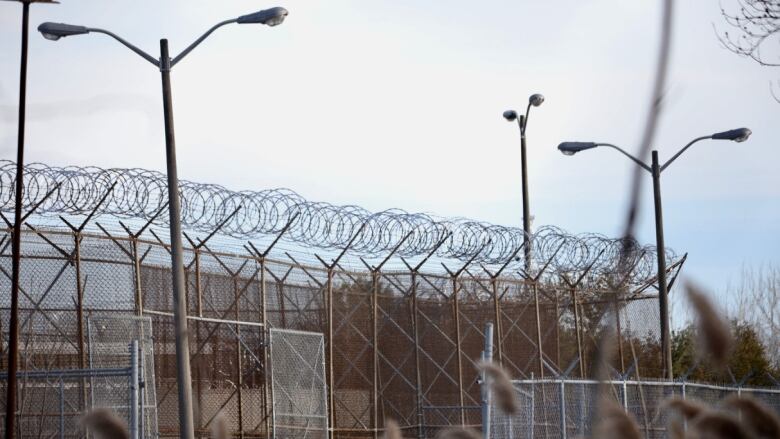 A tall fence topped with barbed wire is seen outside a jail under an overcast sky.