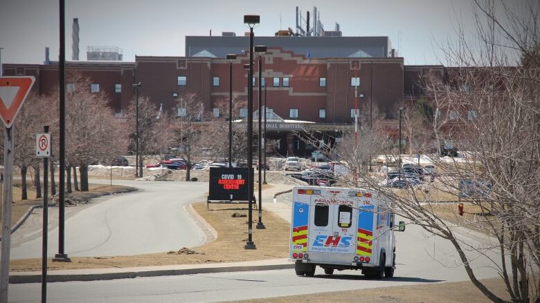 An ambulance drives down a curved road towards a red brick hospital building in the distance.