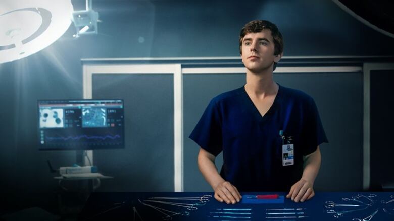 A promotional shot for the medical drama The Good Doctor shows a young, white man in scrubs standing next to a bed in a hospital room with medical instruments laid out in front of him on a steel tray. 