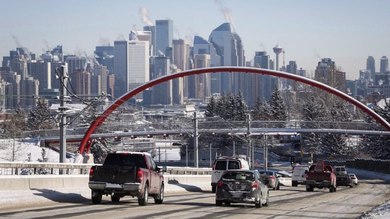 Nearly a dozen dozen cars drive down a road toward downtown Calgary, with the city's skyline in the background. 