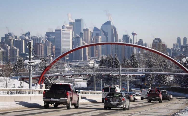 Nearly a dozen dozen cars drive down a road toward downtown Calgary, with the city's skyline in the background. 