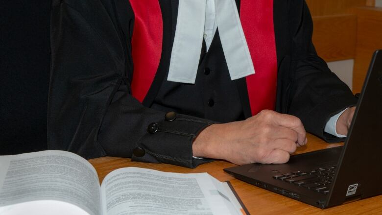 The torso of a judge is shown from below the head. The judge is wearing black robes with red sashes on the shoulders and a white shirt, and they're sitting at a laptop.