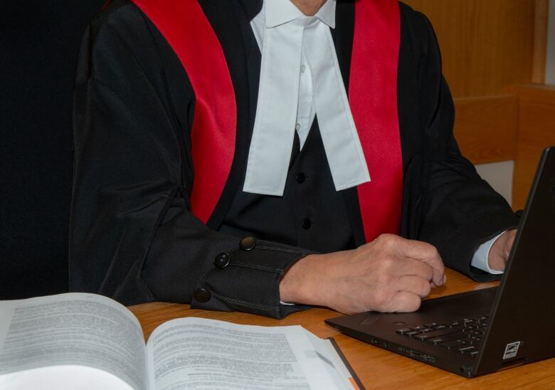 The torso of a judge is shown from below the head. The judge is wearing black robes with red sashes on the shoulders and a white shirt, and they're sitting at a laptop.