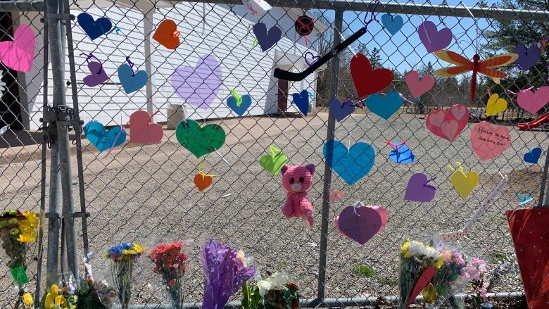 A chain link fence has colourful paper hearts and stuffed animals attached, with flowers and notes on the ground