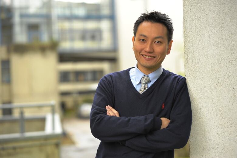 A man wearing a navy coloured sweater over a shirt and tie leans against a wall with his arms crossed.