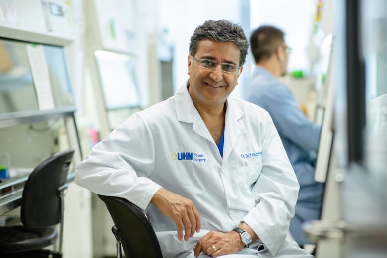 A man in a doctor's coat sits in a hospital, and looks at the camera