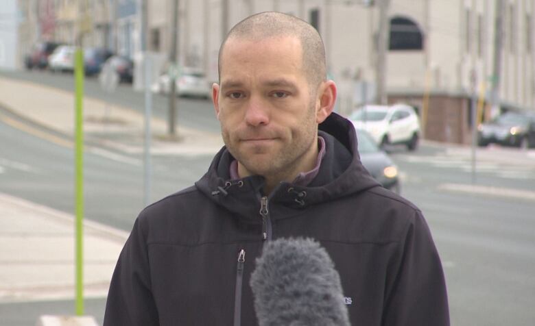 Ian Froude, expressionless, speaking into a microphone in front of City Hall 