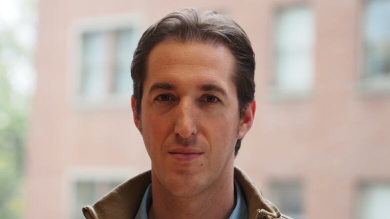 A man with short brown hair slightly smiles for a photo. He's wearing a jacket and a blue button-up shirt.