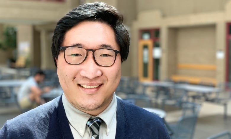 Dr. Jia Hu is standing in an atrium with tables behind him. He's wearing glasses and a navy sweater and is looking directly into the camera.