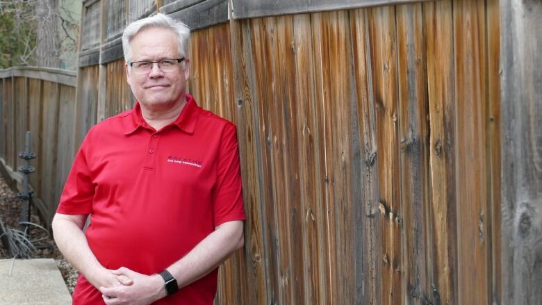 A man with white hair wearing a red shirt leans against a wooden fence.