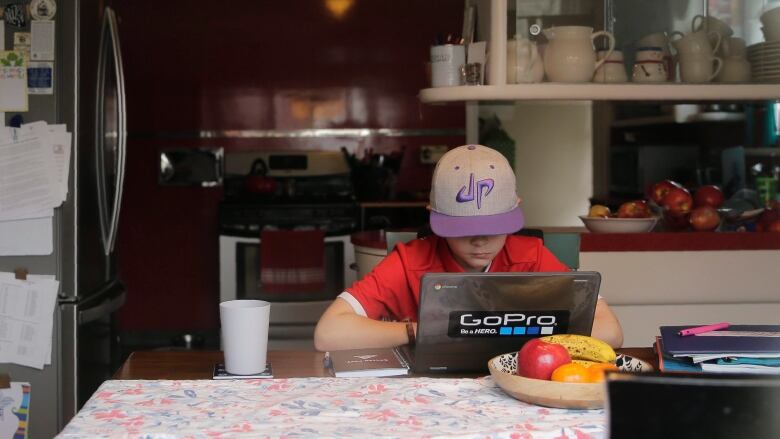 A kid in a red t-shirt and ballcap pulled low over the eyes works at a laptop at a kitchen table. 