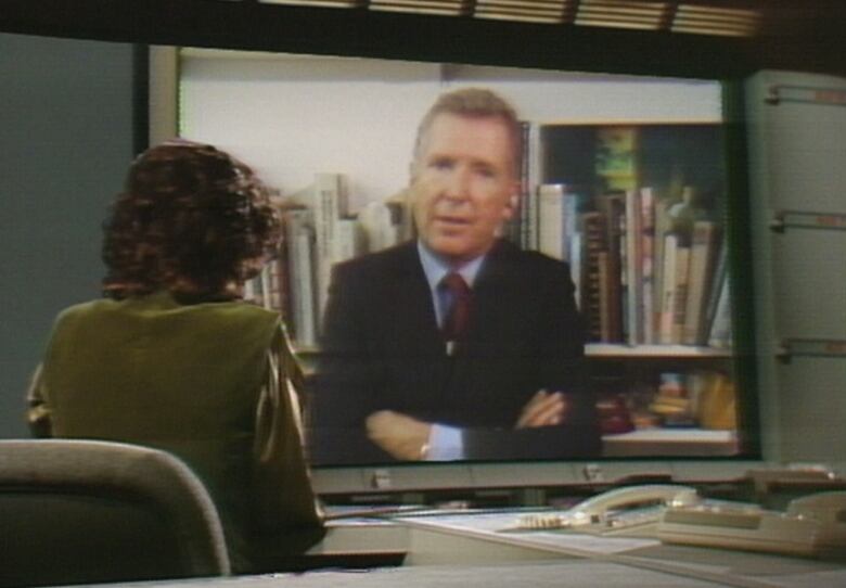 A man on a screen and framed by books speaks to a woman.