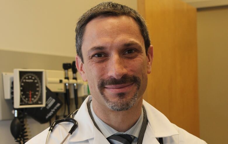 A bearded doctor in a white hospital jacket poses for a photo with a stethoscope around his neck.