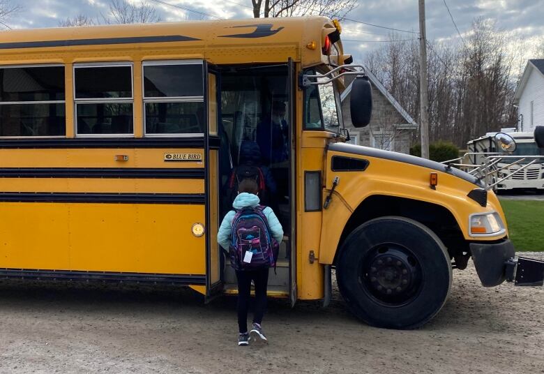 A child in a blue coat and purple backpack boards a yellow school bus 