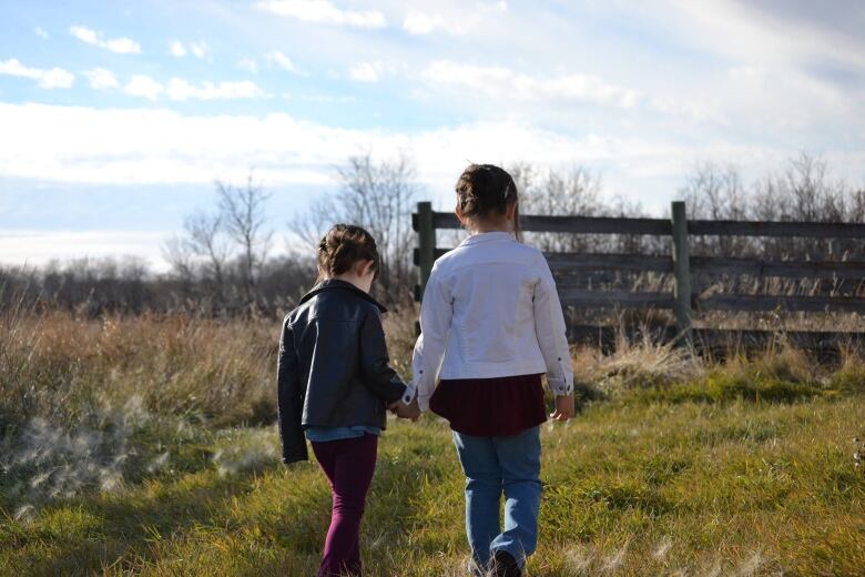 Two young girls hold hands as they walk away from the camera through a farm field on a sunny day.