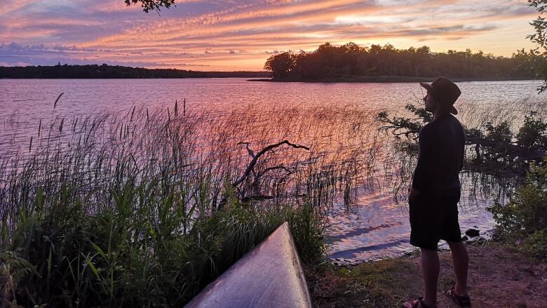 A man stands on a dock by a lake while looking at a sunset. 