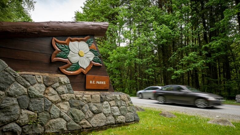 A wooden sign with a dogwood flower that says BC Parks is shown at the entrance to a park while cars pass by on a road surrounded by forest.