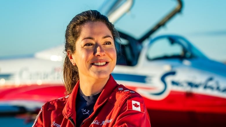 A smiling woman in a red jumpsuit stands in front of a Snowbird training plane.