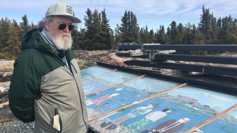 Bearded man stands beside mural lying faceup on gravel