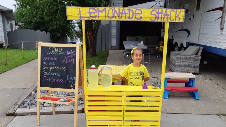 A little girl stands at a lemonade stand in a driveway.