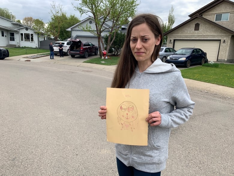 A woman with brown hair stands outside a home with a driveway, holding a piece of paper with a child's drawing.