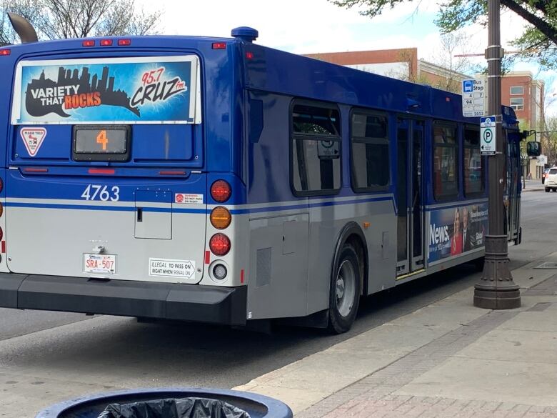 A large blue city bus parks on a busy street. A red brick building, along with traffic lights and other traffic, can be seen in the background. 