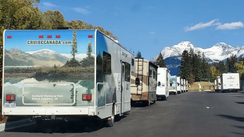 A line of motorhomes can be seen parked on black asphalt with a large mountain in the background surrounded by blue sky.