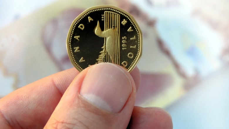 A person holds a loonie with a 20 dollar bill in the background.