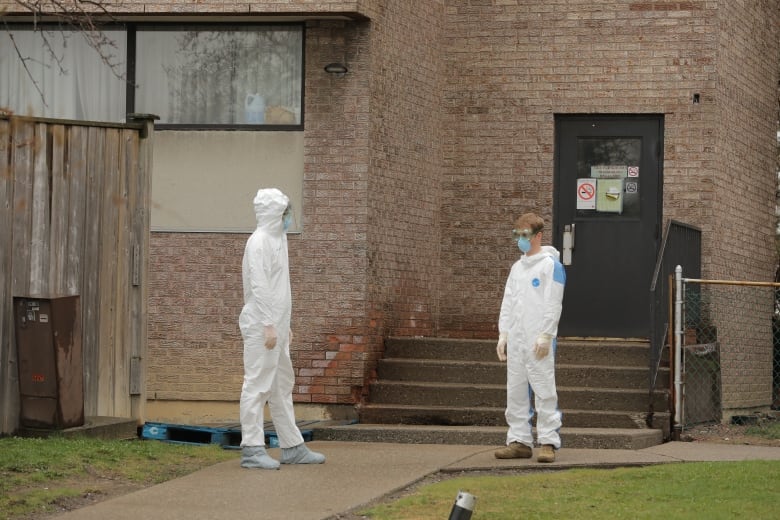 People in white hazmat suits stand outside of a brick building. People in white hazmat suits stand outside of a brick building.