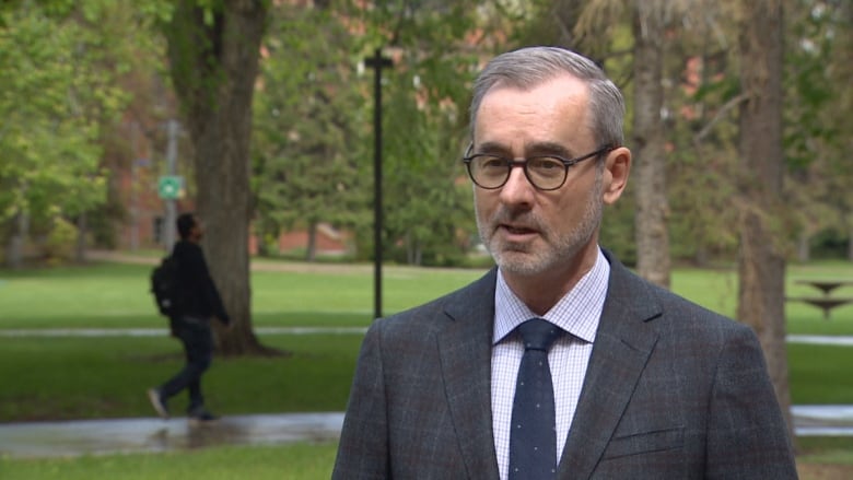 A bearded, bespectacled man stands in front of a park.