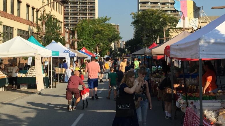 A shot of the downtown Windsor farmers' market is shown on a sunny day with people strolling around and vendors, and their tends, on either side of a street.