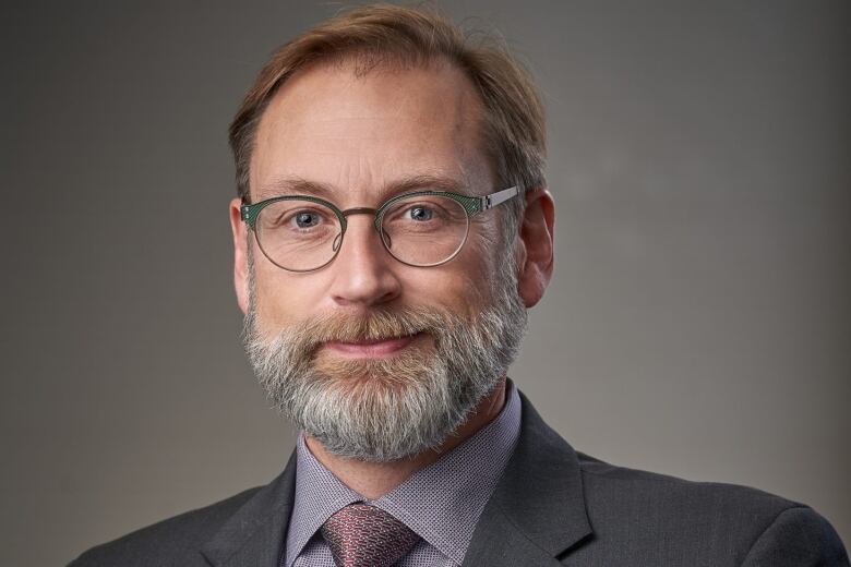 A man with a grey beard and brown hair wearing glasses and a suit poses for a photo 