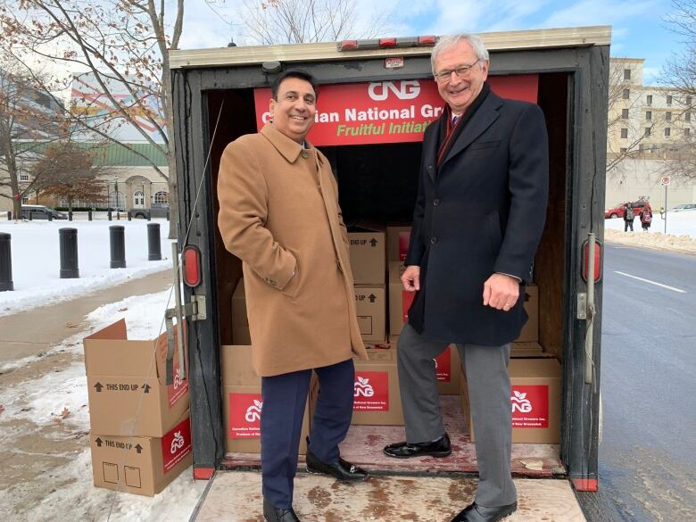 A man in a brown coat on the left with another man in a black coat standing on the ramp of a truck filled with boxes and a sign saying 